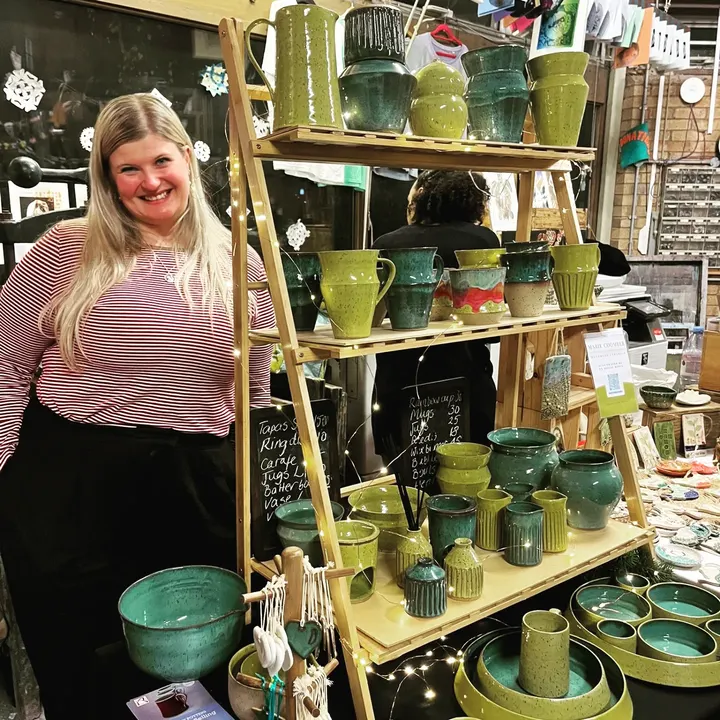 A potter beside shelves of green and teal glazed jugs and vases